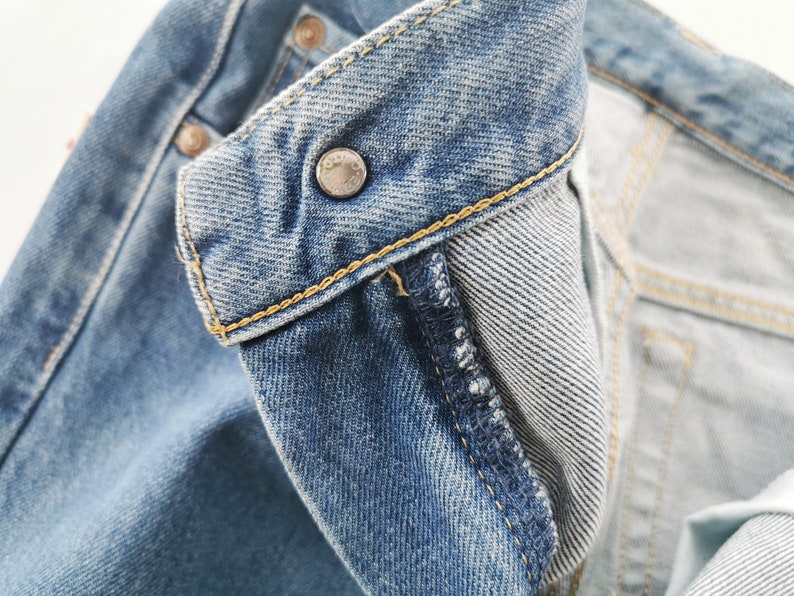 Puede incluir: Primer plano del cuello de una chaqueta vaquera. La chaqueta es de color azul claro con un bot&oacute;n met&aacute;lico y costuras amarillas. El forro interior es de mezclilla azul claro.