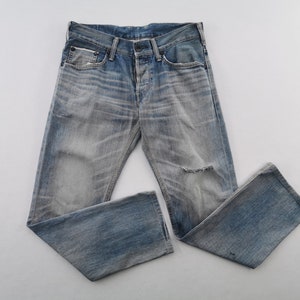 K&ouml;nnte beinhalten: Verblasste blaue Jeans aus Denim mit einem Used-Look, mit Knopfleiste und einem Riss am Knie. Die Jeans liegen flach, wodurch die abgenutzte Textur und die helle Waschung zur Geltung kommen.