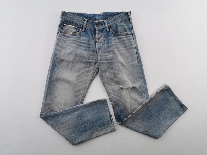 K&ouml;nnte beinhalten: Verblasste blaue Jeans aus Denim mit einem Used-Look, mit Knopfleiste und einem Riss am Knie. Die Jeans liegen flach, wodurch die abgenutzte Textur und die helle Waschung zur Geltung kommen.