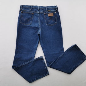 May include: Dark blue denim jeans with a classic five-pocket design. The jeans feature a brown leather patch on the back pocket with the brand name. The jeans are laid flat on a white surface, showcasing the details of the stitching and the Wrangler logo.