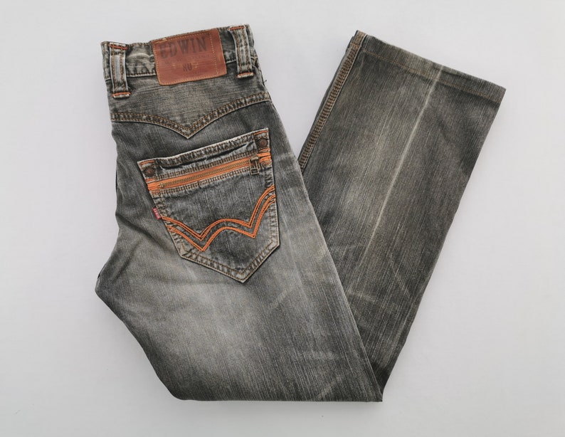 May include: Gray denim jeans with orange stitching and accents. The jeans feature a classic five-pocket design, a leather patch with the brand name "Edwin", and a zip fly. The back pockets have a unique orange stitched design.