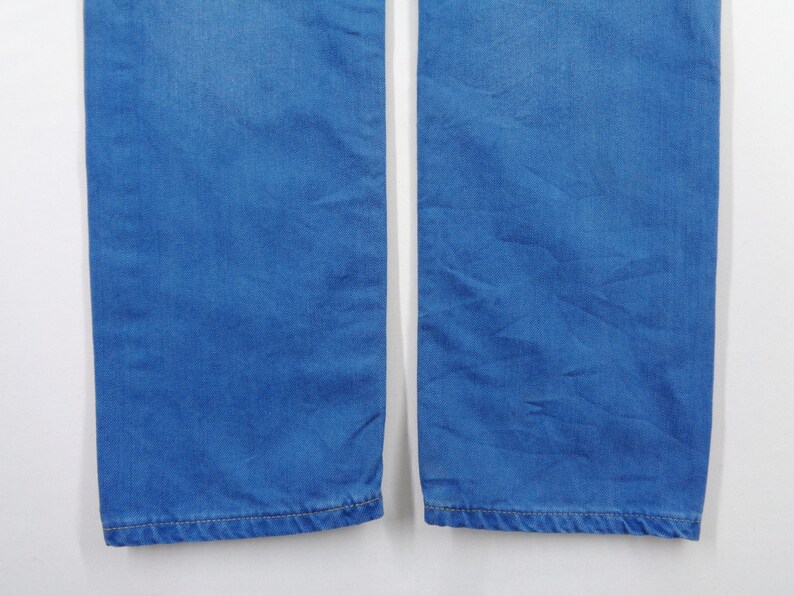 K&ouml;nnte beinhalten: Nahaufnahme des unteren Teils einer blauen Jeans. Der Denim-Stoff hat eine einfarbige, mittelblaue Farbe. Das Bild zeigt die Beine der Jeans mit den S&auml;umen unten. Der Hintergrund ist schlicht wei&szlig;.