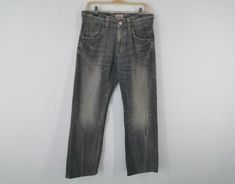 May include: A pair of gray denim jeans hanging on a wooden hanger. The jeans have a faded appearance with visible wear marks and orange stitching. The jeans have a classic five-pocket design and a button and zipper closure.