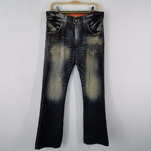Pu&ograve; includere: Jeans in denim blu scuro con un aspetto sbiadito e usurato. I jeans hanno un classico design a cinque tasche, una chiusura con bottoni e cerniera e una leggera apertura della gamba a zampa. I jeans sono appesi a una gruccia di legno.
