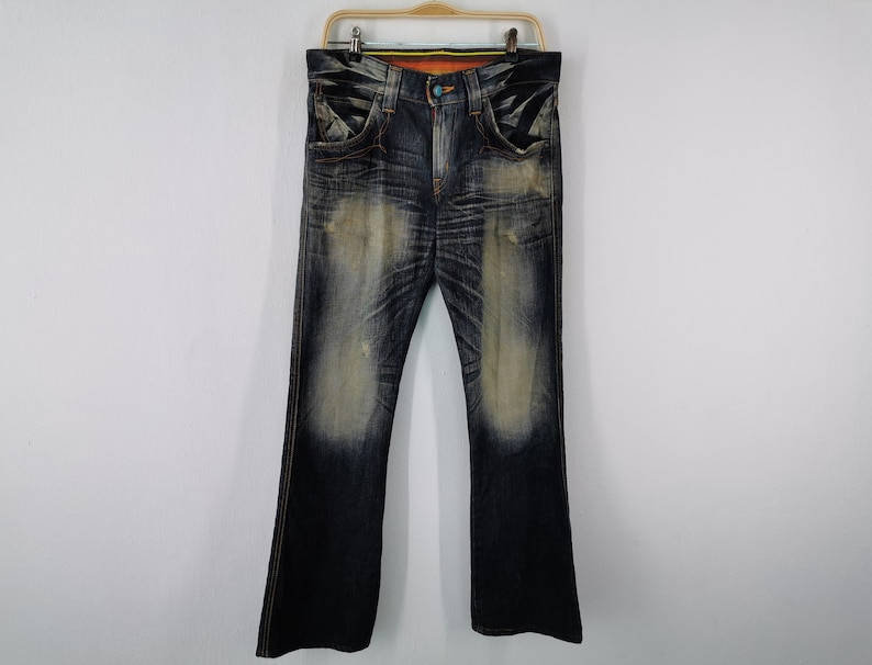Pu&ograve; includere: Jeans in denim blu scuro con un aspetto sbiadito e usurato. I jeans hanno un classico design a cinque tasche, una chiusura con bottoni e cerniera e una leggera apertura della gamba a zampa. I jeans sono appesi a una gruccia di legno.