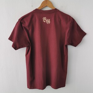 May include: Maroon t-shirt hanging on a wooden hanger. The back of the shirt displays the letters "BG" in a light tan colour. The shirt is made of a soft material and is displayed against a white wall.