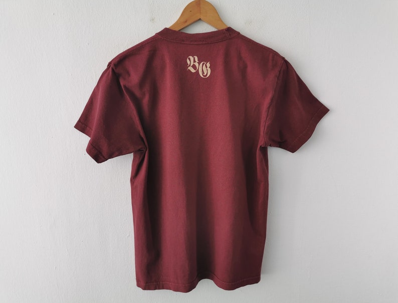 May include: Maroon t-shirt hanging on a wooden hanger. The back of the shirt displays the letters "BG" in a light tan colour. The shirt is made of a soft material and is displayed against a white wall.