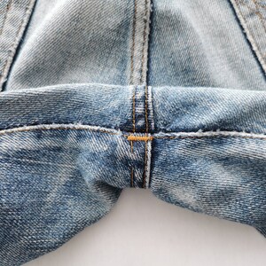 Puede incluir: Primer plano de unos vaqueros de mezclilla azul claro. La imagen destaca las costuras y costuras, con un enfoque en la zona de la entrepierna. El denim tiene un aspecto descolorido y desgastado, con desgaste visible.