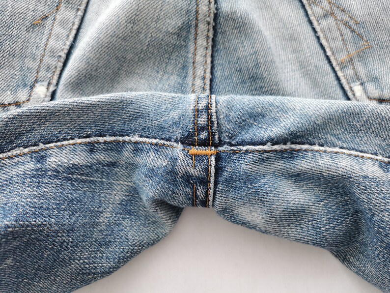 Puede incluir: Primer plano de unos vaqueros de mezclilla azul claro. La imagen destaca las costuras y costuras, con un enfoque en la zona de la entrepierna. El denim tiene un aspecto descolorido y desgastado, con desgaste visible.