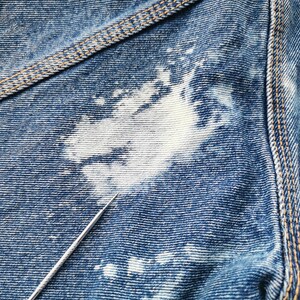 May include: Close-up of blue denim fabric with white bleach spots. The denim has visible texture and stitching in a contrasting tan color. A silver needle points towards the fabric.