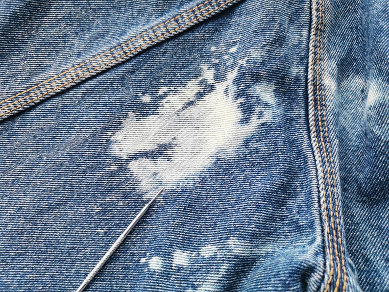 May include: Close-up of blue denim fabric with white bleach spots. The denim has visible texture and stitching in a contrasting tan color. A silver needle points towards the fabric.