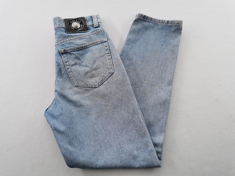 May include: Light blue denim jeans with a classic five-pocket design. Features a leather patch with a logo on the back waistband. The jeans are faded and have a straight leg cut.