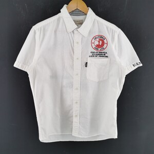 May include: White short-sleeved button-down shirt with a pocket and the text "CALIFORNIA SURF LIFE" in red. The shirt has the text "U.S.N" on the sleeve. The shirt is hanging on a wooden hanger.