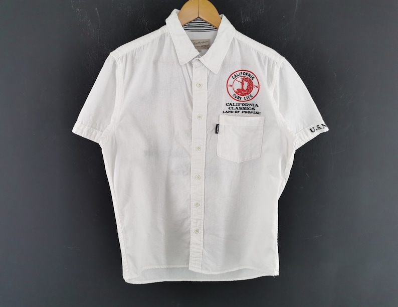 May include: White short-sleeved button-down shirt with a pocket and the text "CALIFORNIA SURF LIFE" in red. The shirt has the text "U.S.N" on the sleeve. The shirt is hanging on a wooden hanger.