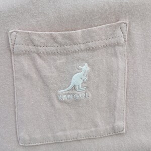 May include: Close-up of a light pink t-shirt pocket with a light blue embroidered kangaroo logo and the brand name "KANGOL". The pocket is rectangular and stitched onto the shirt.