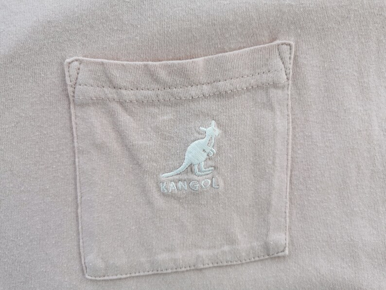 May include: Close-up of a light pink t-shirt pocket with a light blue embroidered kangaroo logo and the brand name "KANGOL". The pocket is rectangular and stitched onto the shirt.