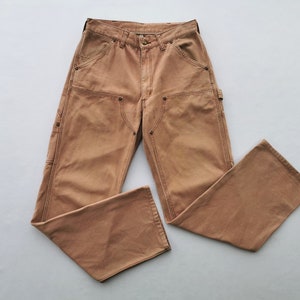 May include: Tan work pants with multiple pockets and reinforced knees. The pants have a button and zipper closure, with a classic five-pocket design. The fabric appears to be a durable cotton blend, suitable for work or casual wear.