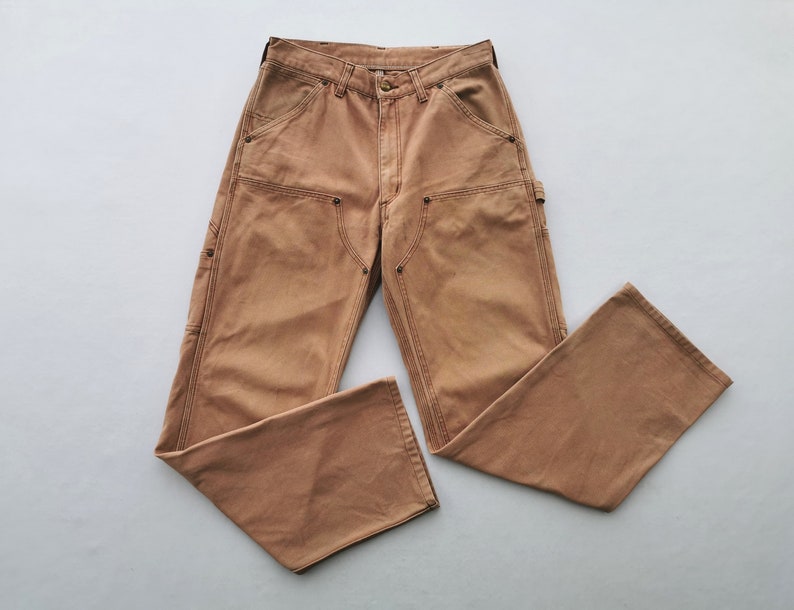 May include: Tan work pants with multiple pockets and reinforced knees. The pants have a button and zipper closure, with a classic five-pocket design. The fabric appears to be a durable cotton blend, suitable for work or casual wear.
