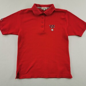 K&ouml;nnte beinhalten: Ein leuchtend rotes Poloshirt mit kurzem &Auml;rmel und Kragen. Es hat eine Knopfleiste mit drei Kn&ouml;pfen und ein gesticktes "COURREGES"-Logo auf der Brust, mit einem kleinen Flaggenlogo auf dem &Auml;rmel. Ein klassisches, l&auml;ssiges Oberteil.