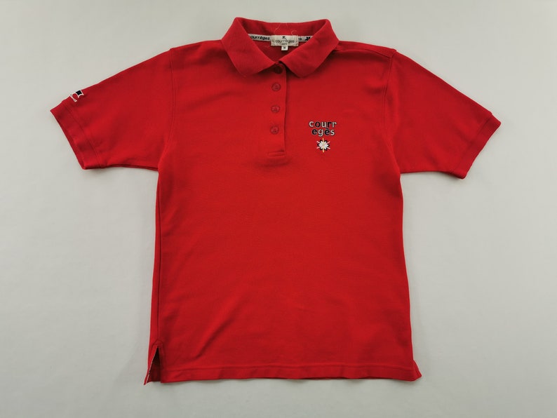 K&ouml;nnte beinhalten: Ein leuchtend rotes Poloshirt mit kurzem &Auml;rmel und Kragen. Es hat eine Knopfleiste mit drei Kn&ouml;pfen und ein gesticktes "COURREGES"-Logo auf der Brust, mit einem kleinen Flaggenlogo auf dem &Auml;rmel. Ein klassisches, l&auml;ssiges Oberteil.