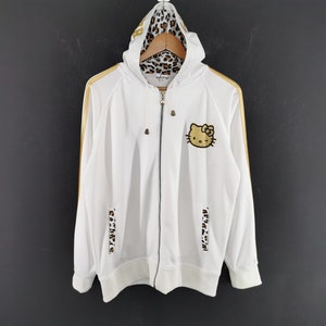May include: White zip-up hoodie with gold stripes and a Hello Kitty patch. The hood lining and lower sleeve accents feature a leopard print design. The hoodie has a drawstring hood and a full-length zipper.