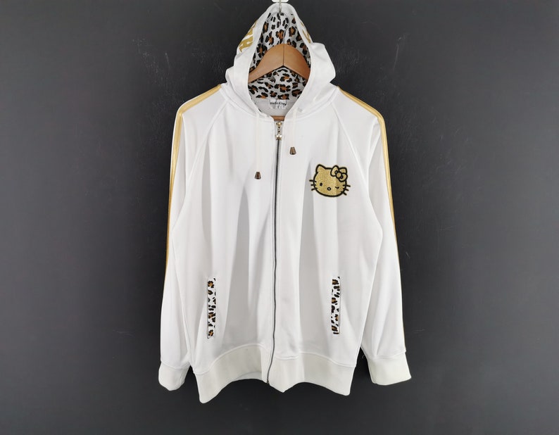 May include: White zip-up hoodie with gold stripes and a Hello Kitty patch. The hood lining and lower sleeve accents feature a leopard print design. The hoodie has a drawstring hood and a full-length zipper.
