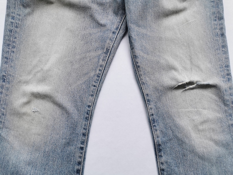 May include: Light blue denim jeans with distressed details, including a tear on the right knee. The jeans have a faded wash and visible stitching along the seams. The image shows the upper portion of the jeans.
