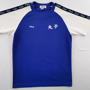 Puede incluir: Una camiseta deportiva azul y blanca con la marca "FILA" en el pecho y las mangas. Las mangas son blancas con "FILA" impreso en negro. Caracteres japoneses est&aacute;n impresos en la parte delantera.