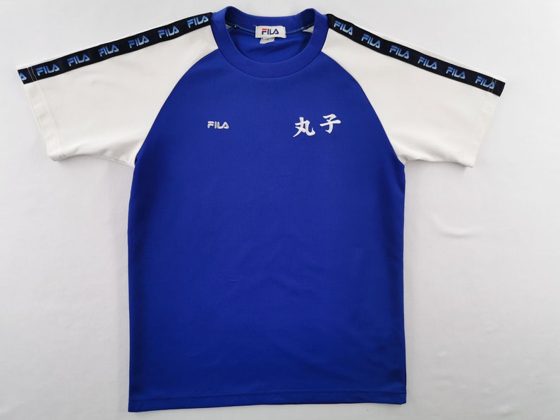 Puede incluir: Una camiseta deportiva azul y blanca con la marca "FILA" en el pecho y las mangas. Las mangas son blancas con "FILA" impreso en negro. Caracteres japoneses est&aacute;n impresos en la parte delantera.