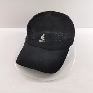 Puede incluir: Gorra de b&eacute;isbol Kangol negra con visera curva. La gorra presenta un logotipo de canguro blanco y el nombre de la marca "KANGOL" en la parte delantera. La parte superior de la gorra tiene un dise&ntilde;o de malla. La gorra est&aacute; sobre un plato transparente.