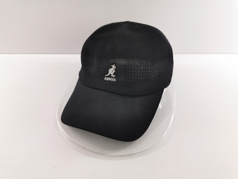 Puede incluir: Gorra de b&eacute;isbol Kangol negra con visera curva. La gorra presenta un logotipo de canguro blanco y el nombre de la marca "KANGOL" en la parte delantera. La parte superior de la gorra tiene un dise&ntilde;o de malla. La gorra est&aacute; sobre un plato transparente.