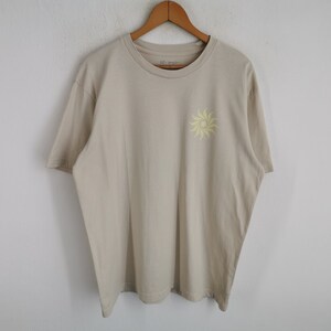 May include: A beige short-sleeved t-shirt featuring a crew neckline and a small, pale yellow sun graphic printed on the upper left chest area. The garment is displayed on a wooden hanger, showcasing a simple design with a solid neutral colour.