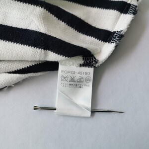 Puede incluir: Primer plano de una tela a rayas blancas y azul marino. La tela est&aacute; doblada, revelando una etiqueta con s&iacute;mbolos de cuidado y el texto "ECPGI-45190". Un alfiler plateado est&aacute; colocado en la etiqueta.