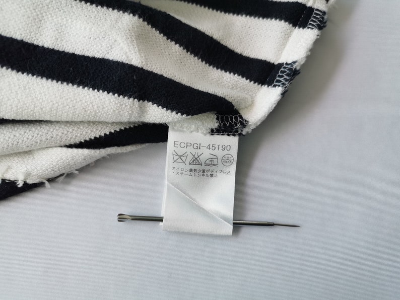 Puede incluir: Primer plano de una tela a rayas blancas y azul marino. La tela est&aacute; doblada, revelando una etiqueta con s&iacute;mbolos de cuidado y el texto "ECPGI-45190". Un alfiler plateado est&aacute; colocado en la etiqueta.