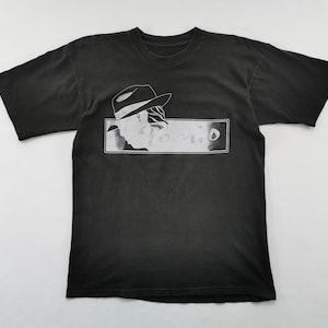May include: Black t-shirt featuring a white graphic of a person in a fedora hat and the word "Sonic" in a rectangular frame. The shirt is made of a soft material.