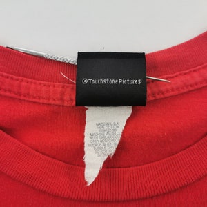 May include: Close-up of a red t-shirt collar with a black tag that reads "&copy; Touchstone Pictures." Below the tag is a white label with text including "MADE IN U.S.A." and "100% COTTON."