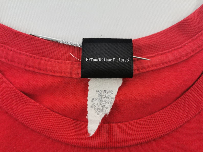 May include: Close-up of a red t-shirt collar with a black tag that reads "&copy; Touchstone Pictures." Below the tag is a white label with text including "MADE IN U.S.A." and "100% COTTON."