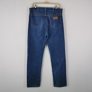 May include: A pair of dark blue denim jeans featuring a classic five-pocket design, a high-rise waist, and a straight-leg cut. The back view displays a brown leather patch on the right pocket, copper-toned rivets, and visible stitching details.