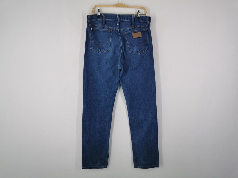 May include: A pair of dark blue denim jeans featuring a classic five-pocket design, a high-rise waist, and a straight-leg cut. The back view displays a brown leather patch on the right pocket, copper-toned rivets, and visible stitching details.