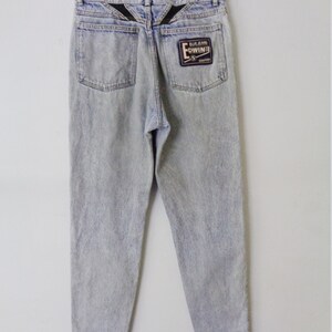 May include: Light blue acid wash jeans with a faded appearance, hanging on a hanger. The jeans feature two back pockets and a black patch with the brand name "EDWIN" on the right pocket. The back yoke has a black V-shaped design.