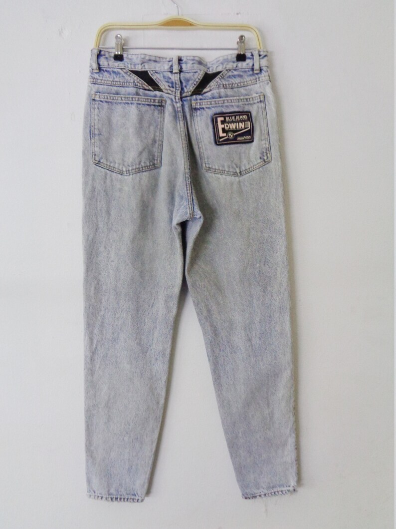 May include: Light blue acid wash jeans with a faded appearance, hanging on a hanger. The jeans feature two back pockets and a black patch with the brand name "EDWIN" on the right pocket. The back yoke has a black V-shaped design.