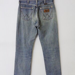 Pu&ograve; includere: Jeans di denim blu sbiadito con il logo Wrangler sulla tasca posteriore. I jeans sono appesi a una gruccia su uno sfondo bianco. I jeans hanno un classico design a cinque tasche e una gamba dritta.