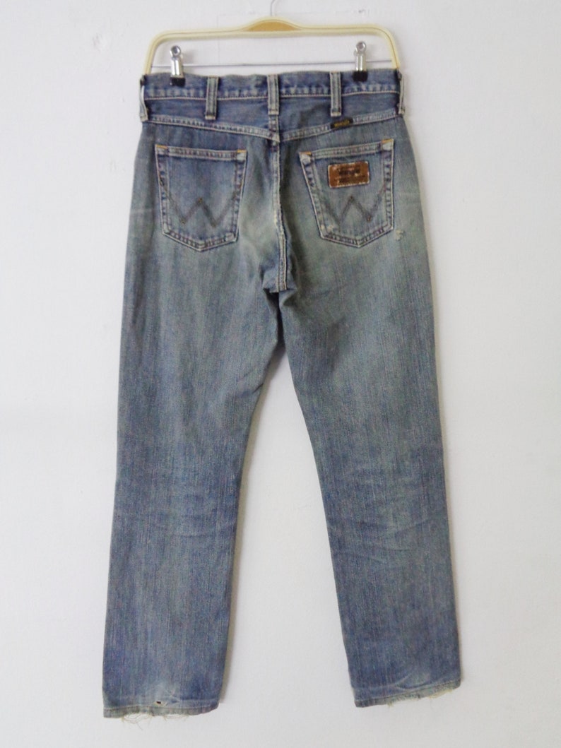 Pu&ograve; includere: Jeans di denim blu sbiadito con il logo Wrangler sulla tasca posteriore. I jeans sono appesi a una gruccia su uno sfondo bianco. I jeans hanno un classico design a cinque tasche e una gamba dritta.