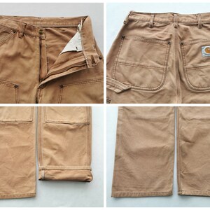 May include: Close-up shots of tan work pants. The pants have multiple pockets, reinforced stitching, and a button and zipper closure. The Carhartt logo is visible on the back pocket. The cuffs are folded up on one leg.