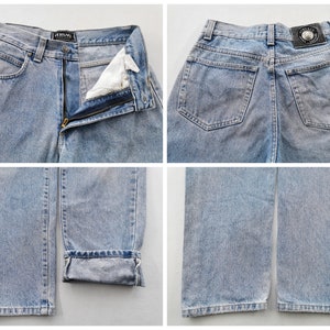 May include: Light blue denim jeans with a classic five-pocket design. The jeans feature a zip fly, belt loops, and a faded wash. The Versace logo is visible on the waistband and back patch. The jeans are cuffed at the hem.
