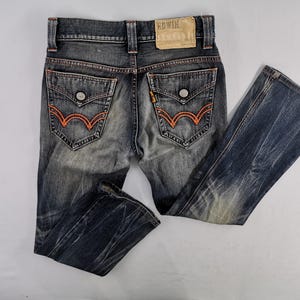 Op de afbeelding: Verbleekte blauwe denim jeans met oranje stiksels. Voorzien van twee achterzakken met oranje borduursel en knoopsluitingen. De jeans heeft een bootcut beenopening en een leren patch met de merknaam "EDWIN".