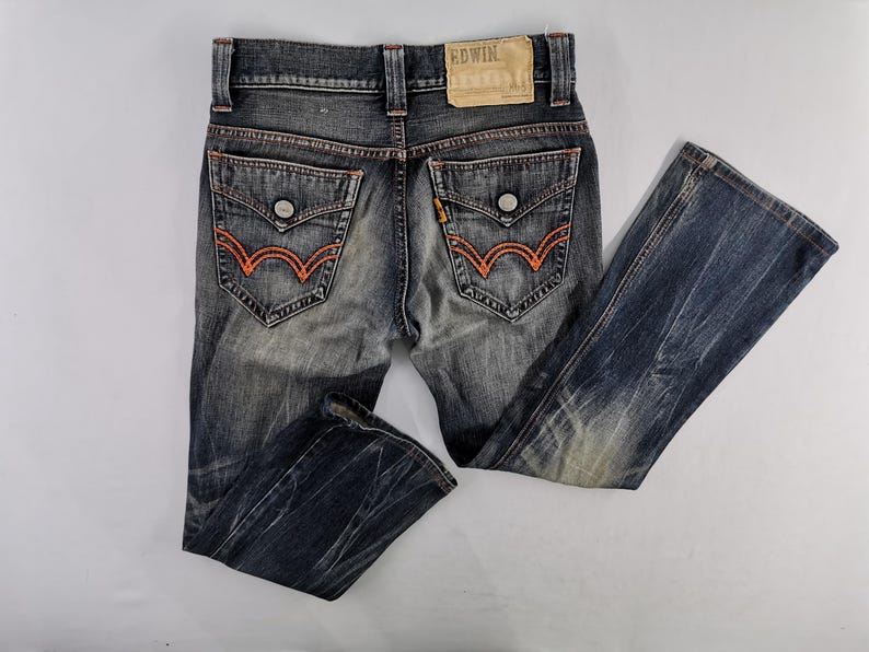 Op de afbeelding: Verbleekte blauwe denim jeans met oranje stiksels. Voorzien van twee achterzakken met oranje borduursel en knoopsluitingen. De jeans heeft een bootcut beenopening en een leren patch met de merknaam "EDWIN".