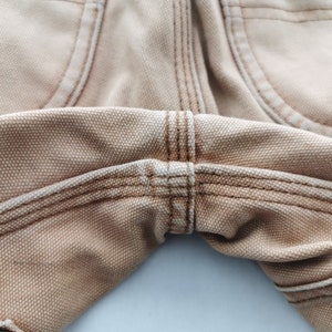 May include: Close-up of tan-colored work pants, showcasing the detailed stitching and fabric texture. The image highlights the seams and pocket details, emphasizing the durable construction and design of the garment. The pants appear to be made of a sturdy canvas material.