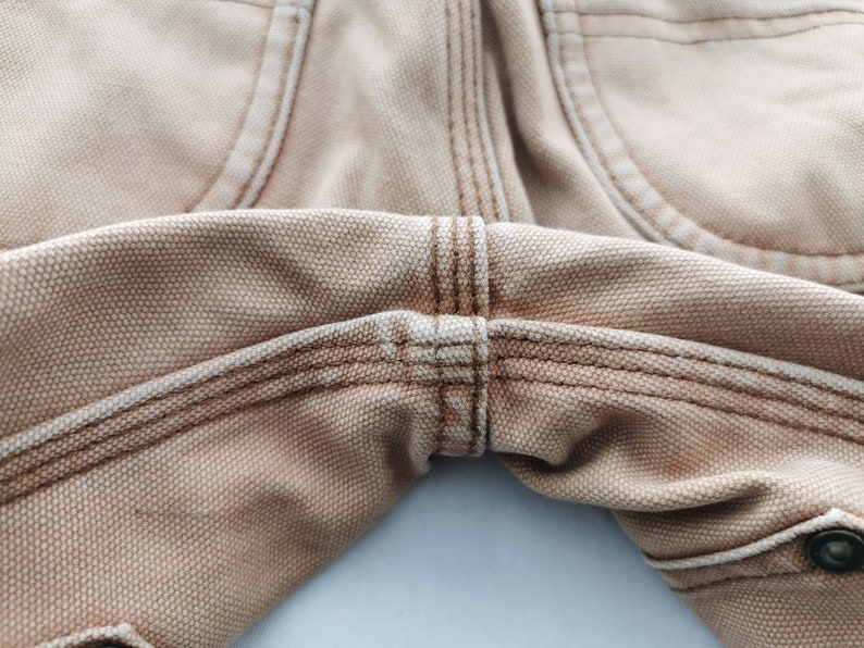May include: Close-up of tan-colored work pants, showcasing the detailed stitching and fabric texture. The image highlights the seams and pocket details, emphasizing the durable construction and design of the garment. The pants appear to be made of a sturdy canvas material.