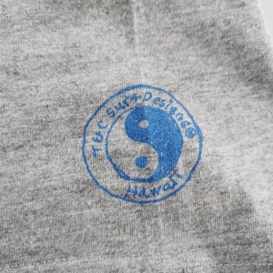 May include: Close-up of a gray t-shirt with a blue circular logo. The logo features a yin-yang symbol and the text "T&C Surf Designs Hawaii". The fabric has a heathered texture.